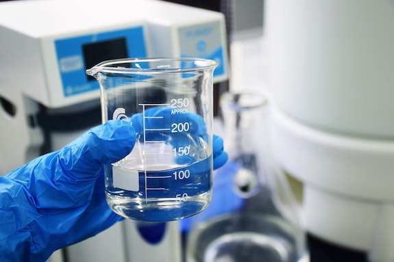 A blue-gloved hand holding a 250 mL beaker filled with a clear, colorless solution.