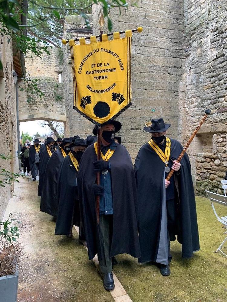 A parade of people wearing black and holding a yellow pendant with a truffle on it. The pendant reads 'Conférie du diamant noir et de la gastronomie tuber melanosporum'